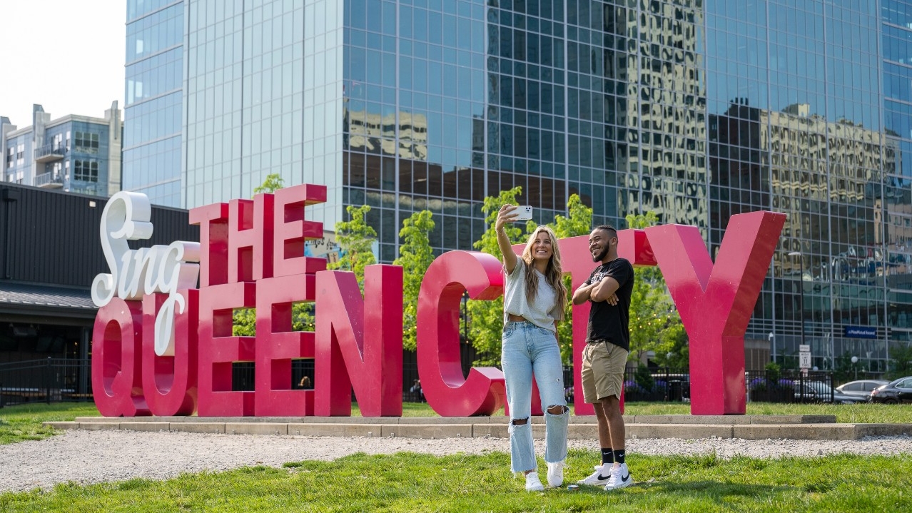 Two students taking a photo next to a sign reading "sing the queen city"